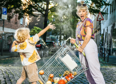 Marie Lumpp mit Kind und Einkaufswagen auf der Straße. Das Kind bewirft sie mit Mehl