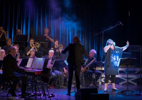 Sängerin steht auf der Bühne begleitet von einem Orchester